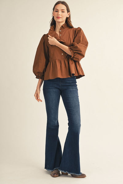 A woman modeling a brown ruffled long-sleeve blouse with puff sleeves and dark blue flared jeans, standing against a plain light beige studio background.