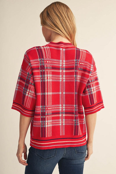 A woman viewed from the back wearing a red plaid knit sweater with white and navy lines, paired with blue jeans, against a plain beige background.