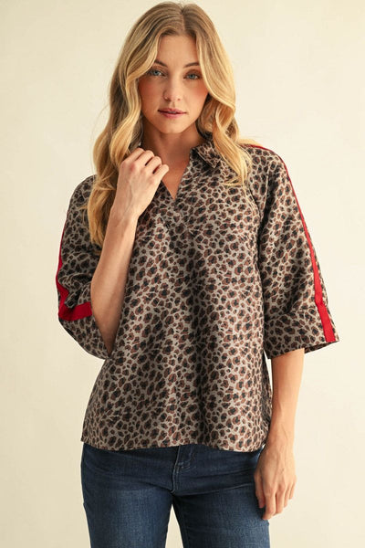 A woman modeling a leopard print short-sleeve blouse with red stripe accents on the sleeves, paired with blue jeans, against a plain beige background.