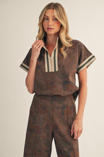 A woman modeling a brown short-sleeve top with a floral jacquard pattern, featuring a split V-neckline with cream and dark green stripes, paired with matching pants against a light beige background.