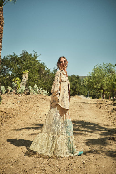 Woman in a long, light-colored dress standing in a desert landscape with cacti and palm trees.