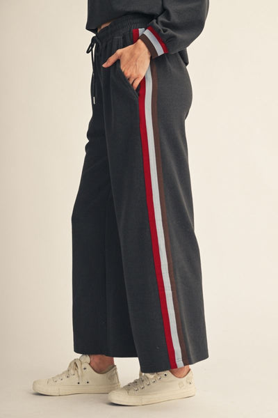 A person wearing dark charcoal wide-leg sweatpants with a red, white, and gray side stripe, paired with white sneakers, shown from the waist down against a light beige studio background.
