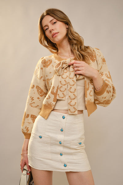 Woman wearing a patterned beige cardigan over a beige top with a white skirt featuring blue buttons on a plain background