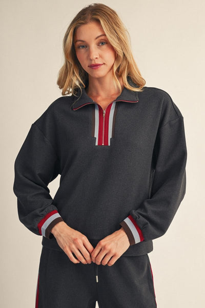 A woman modeling a dark charcoal gray quarter-zip sweatshirt with red and white striped trim on the collar and cuffs, paired with matching dark sweatpants against a light beige studio background.