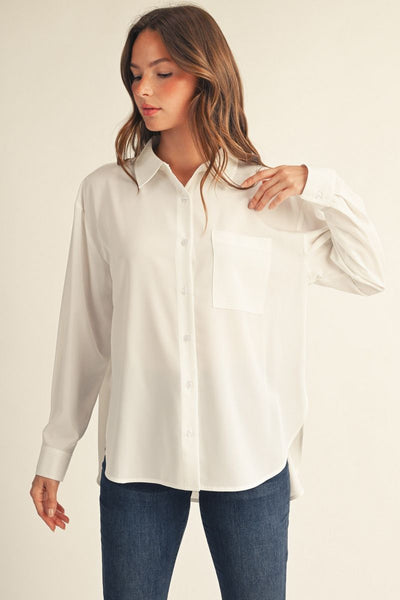 A woman wearing a white long-sleeve button-up shirt with a chest pocket, paired with blue jeans, photographed against a plain light beige studio background.