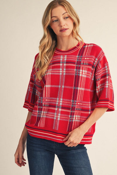A woman wearing a red plaid short-sleeve knit top with blue jeans, photographed against a plain beige studio background.