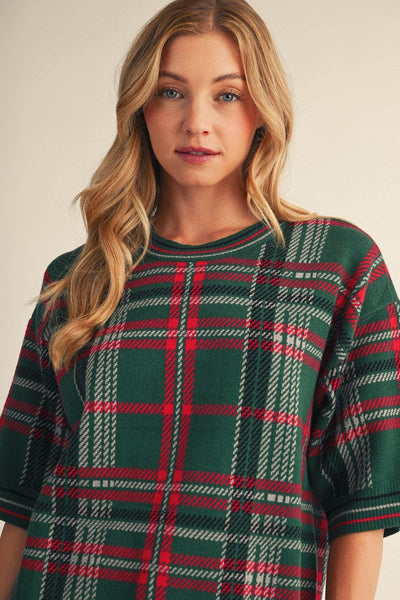 A woman wearing a green plaid short-sleeve top with red and white intersecting stripes, photographed against a plain light beige studio background.