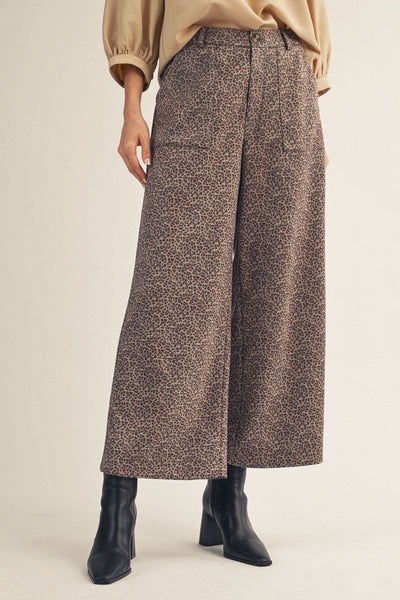 A person wearing high-waisted, wide-leg cropped trousers with an allover brown and beige animal print, styled with a beige top and black ankle boots against a plain beige studio background.