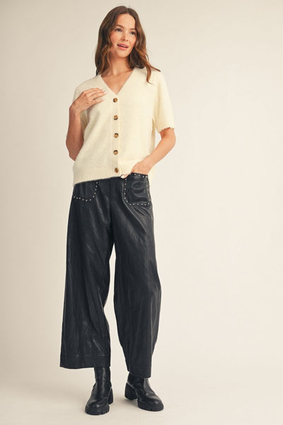 A woman modeling a cream short-sleeve knit cardigan with brown buttons, paired with black wide-leg pants and black ankle boots, against a plain beige studio background.