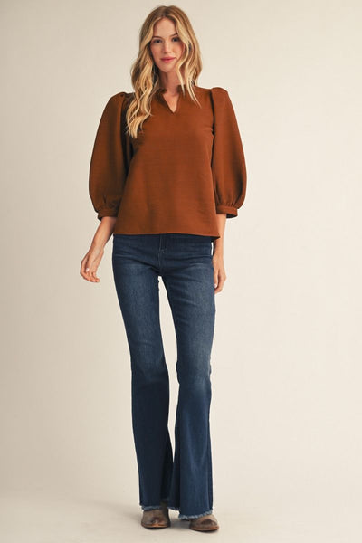 A woman modeling a rust-brown long-sleeve blouse with puffed sleeves and a split V-neck, paired with dark blue flare jeans and brown shoes, against a plain beige studio background.