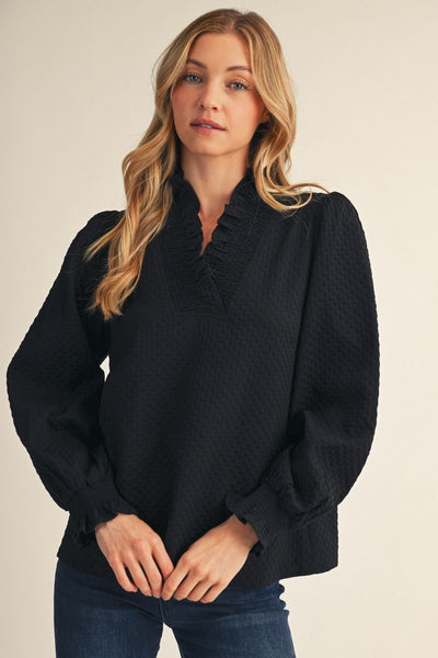 A woman modeling a black textured long-sleeve blouse with a ruffled split neckline, paired with blue jeans, against a plain beige studio background.