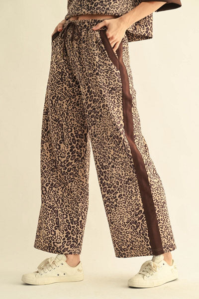 A person wearing wide-leg leopard-print pants with a dark brown side stripe, paired with white sneakers, shown from the waist down against a plain light beige studio background.