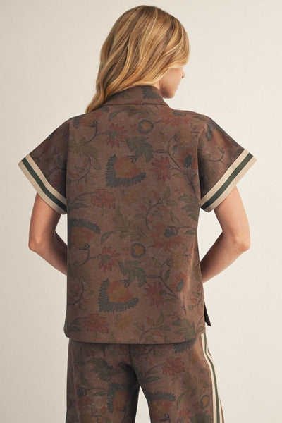 Back of a model wearing a brown short-sleeve top with a floral jacquard pattern, featuring a split V-neckline with cream and dark green stripes, paired with matching pants against a light beige background.