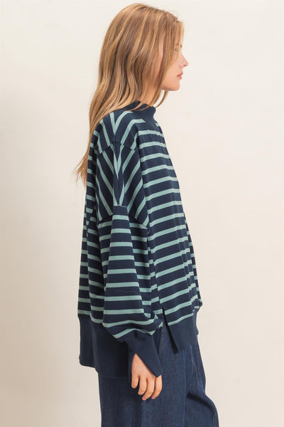 side view of a woman wearing a navy/sage striped sweatshirt and jeans with a light background