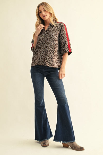 A woman modeling a leopard-print button-up shirt with red and black striped sleeves, paired with dark blue flare jeans and brown boots, against a plain beige studio background.