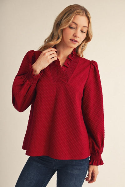 A woman wearing a textured red long-sleeve blouse with a ruffled V-neckline and puff sleeves, paired with dark blue jeans, against a plain beige background.