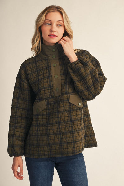 A woman wearing a quilted plaid jacket in olive green and black, paired with blue jeans, standing against a plain beige studio background.