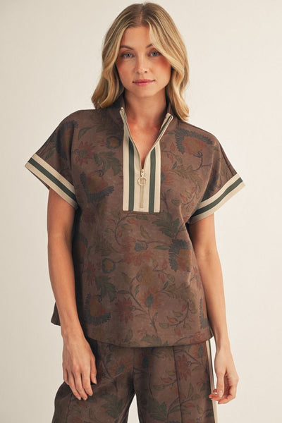 A woman modeling a brown short-sleeve top with a floral jacquard pattern, featuring a split V-neckline with cream and dark green stripes, paired with matching pants against a light beige background.