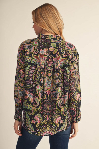 A woman shown from the back wearing a long-sleeve high-neck blouse with an intricate multicolor paisley and floral pattern on a black base, paired with dark blue jeans, against a plain beige background.