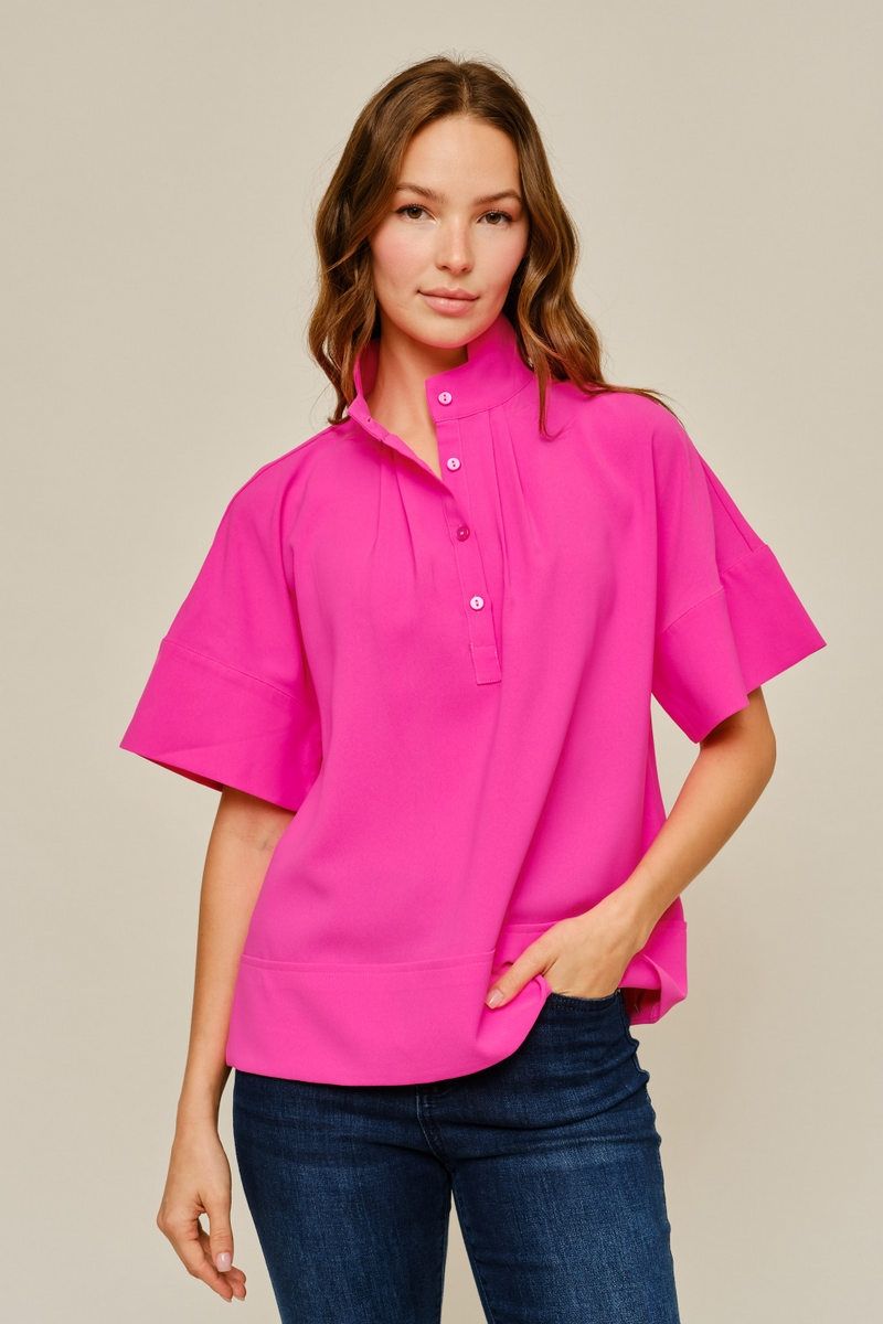 A woman modeling a bright pink short-sleeve button-up shirt with a mock neck, paired with dark blue jeans, against a solid light beige background.