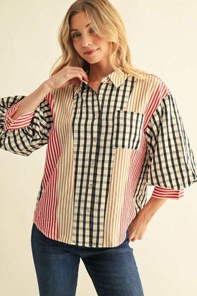 A woman wearing a patchwork button-up shirt with vertical stripes, gingham, and solid panels in beige, black, and red, paired with dark blue jeans against a light beige background.