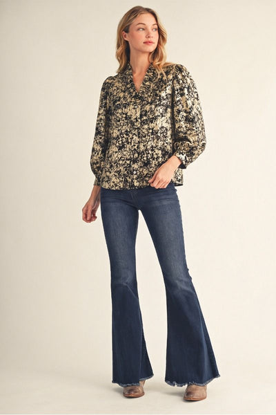 A woman wearing a long-sleeve button-up blouse with a black and beige abstract floral pattern, paired with dark blue jeans, against a plain beige background.