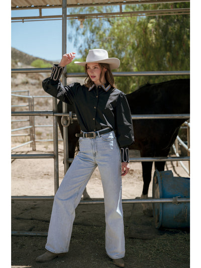Person wearing a black leather jacket, white cowboy hat, and light blue jeans standing in a stable with a horse.