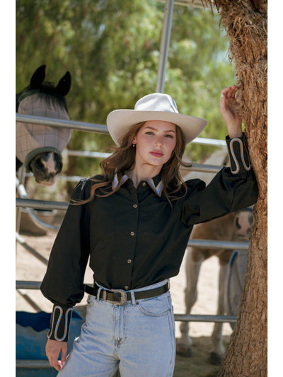 Woman in a black shirt and white cowboy hat standing next to a horse in a corral.