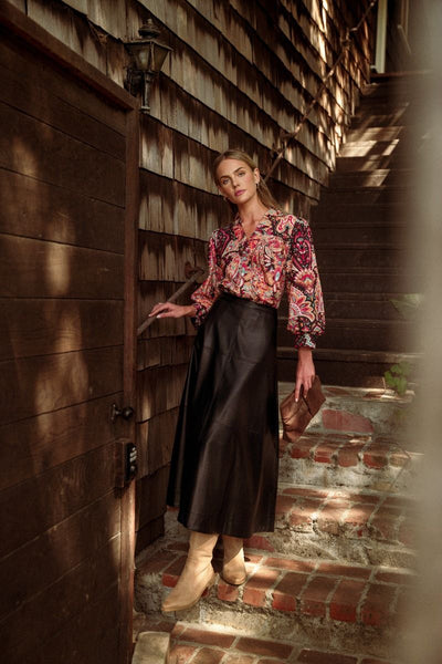 A woman stands on outdoor brick stairs beside a rustic wooden building, wearing a long-sleeve floral blouse with a dark midi skirt and tan ankle boots, while holding a brown clutch.