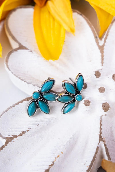 A pair of silver post earrings with blue stone accents, designed in a floral pattern, displayed on a white surface with a yellow flower in the background.
